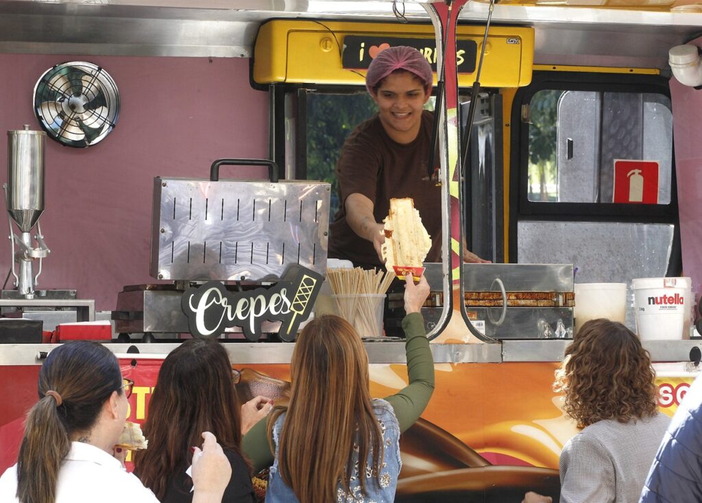 A imagem mostra um food truck especializado em crepes, com uma comerciante usando touca e avental preparando e entregando um crepe a uma cliente. Ao redor do veículo, outras pessoas aguardam atendimento. O espaço de preparo conta com equipamentos de cozinha, utensílios e ingredientes, incluindo um pote de Nutella. O ambiente transmite a movimentação de um evento gastronômico ao ar livre.