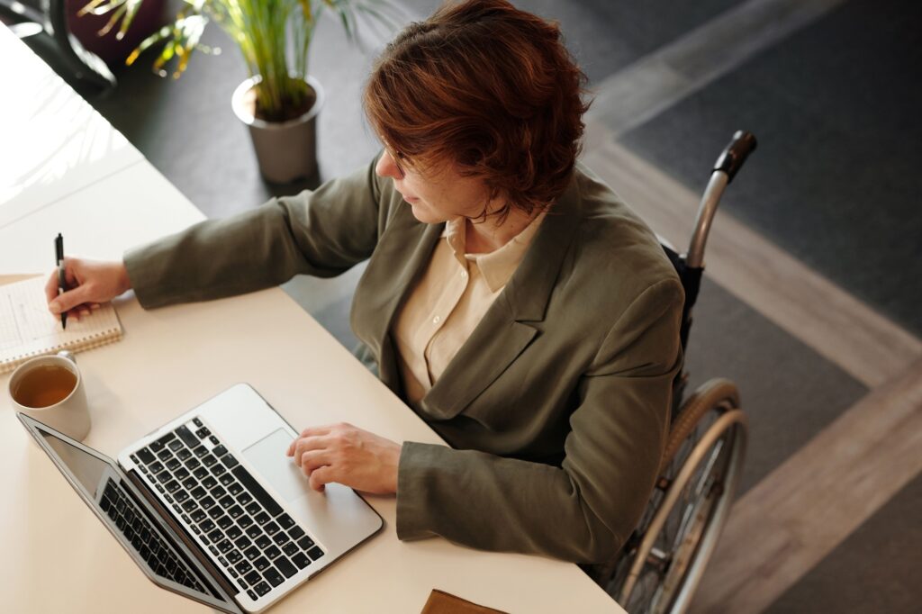 A imagem mostra uma mulher sentada em uma cadeira de rodas, trabalhando em uma mesa. Ela escreve em um caderno com uma mão e usa um laptop com a outra. Ao lado, há uma xícara de bebida quente e, ao fundo, uma planta em vaso. O ambiente é moderno, bem iluminado e parece ser um escritório ou espaço de estudo.