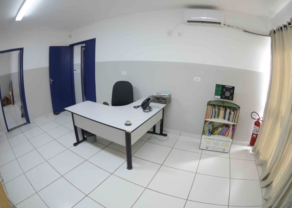 A imagem mostra um escritório simples e bem organizado, com piso branco e paredes em dois tons — cinza claro e branco. Há uma mesa de trabalho com telefone, pilhas de documentos e uma campainha, além de uma cadeira preta posicionada atrás dela. No canto direito, há uma estante com livros e um cartaz que diz “Tambor Literário — Pegue um, Deixe Outro”, incentivando a troca de livros. Um extintor de incêndio e cortinas claras completam o ambiente, que transmite uma sensação de limpeza e funcionalidade.