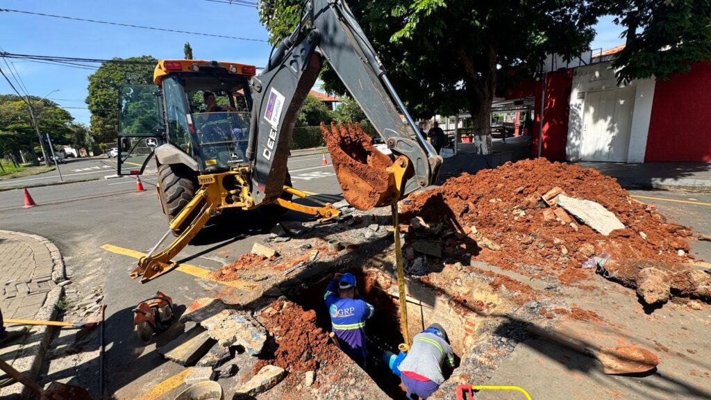 Uma retroescavadeira amarela da John Deere está em operação em uma vala aberta na rua, removendo terra com sua pá. Dois trabalhadores em roupas de trabalho estão dentro da vala, um deles parece estar ajustando uma tubulação azul. A terra removida está empilhada em um monte em primeiro plano e ao lado da escavação. A cena ocorre em uma rua pavimentada, com um prédio vermelho e branco ao fundo. Árvores fornecem sombra parcial. O céu está claro e azul. A composição foca na atividade de construção, destacando a maquinaria e o trabalho manual. As cores predominantes são o amarelo da retroescavadeira, o marrom da terra, o azul do céu e das roupas de trabalho, e o vermelho do prédio.