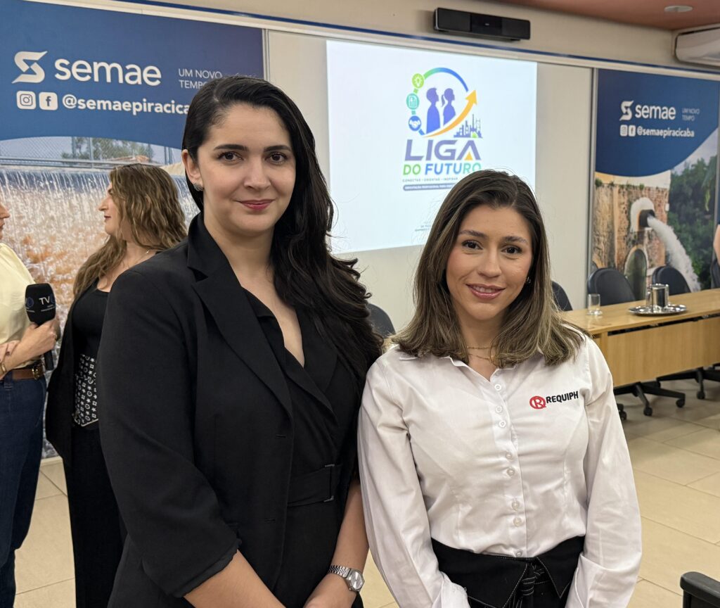 A imagem mostra duas mulheres em destaque, sorrindo e posando para a foto em um ambiente corporativo. Ao fundo, há um telão com o logotipo do projeto “Liga do Futuro”, iniciativa da Prefeitura de Piracicaba voltada à orientação profissional e desenvolvimento de carreira, além de banners do SEMAE Piracicaba, indicando parceria institucional.