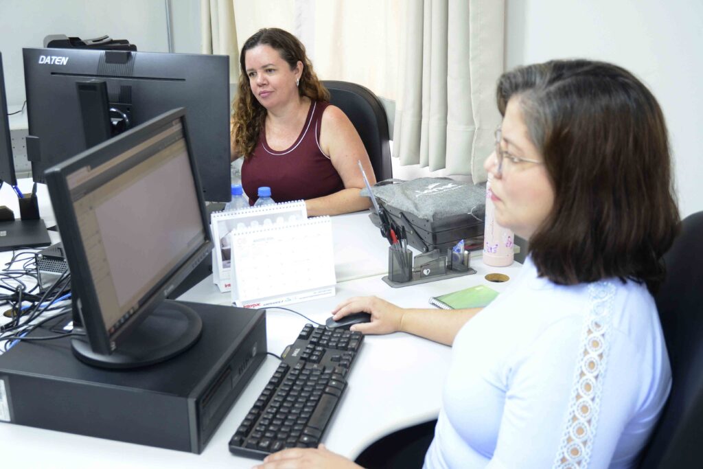 A imagem mostra duas mulheres trabalhando em um escritório, cada uma diante de um computador de mesa