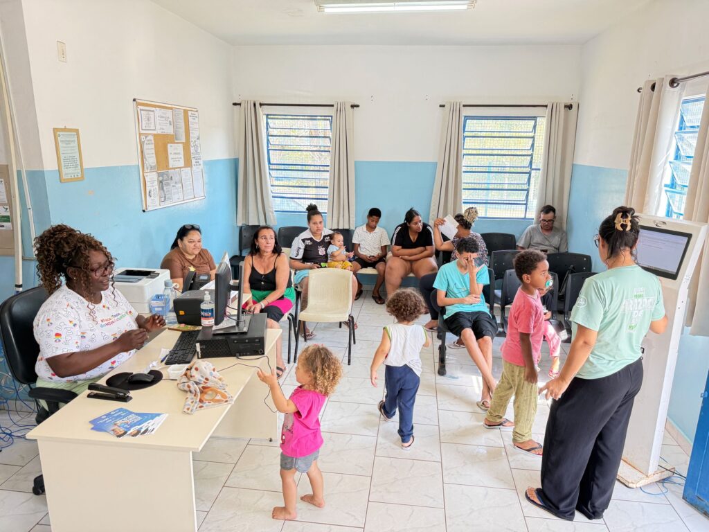 A imagem mostra uma sala de atendimento ou serviço público, com paredes brancas e azuis, janelas com grades e cortinas claras. Há várias pessoas sentadas em cadeiras, aguardando atendimento, incluindo adultos, crianças e um bebê. No lado esquerdo, uma funcionária trabalha diante de um computador, impressora e outros equipamentos. À direita, outra profissional, usando camiseta verde-clara, auxilia uma pessoa em um terminal de autoatendimento. Um quadro de avisos com cartazes e comunicados está fixado na parede.