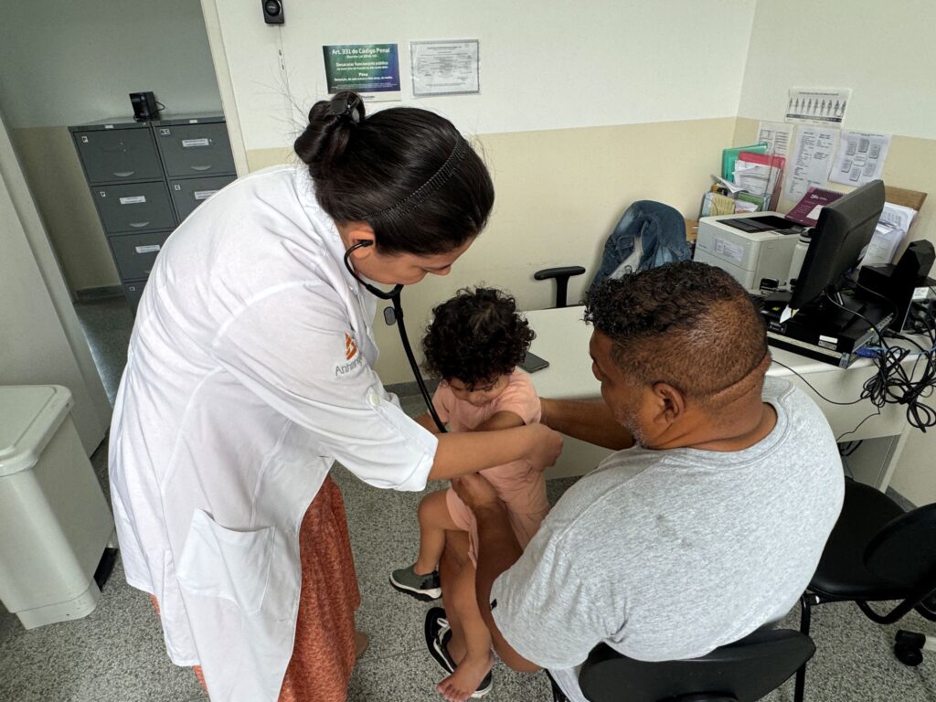 A imagem mostra uma profissional da área da saúde examinando uma criança pequena com um estetoscópio. Ela usa jaleco branco, prende o cabelo e se inclina para auscultar o peito da criança, que está no colo de um homem. O homem, sentado em uma cadeira, segura a criança com cuidado. O atendimento acontece em uma sala de serviço de saúde, com mesa, computador, impressora, armários para documentos e cartazes informativos nas paredes.
