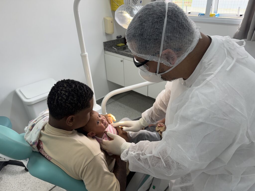 A imagem mostra um atendimento odontológico infantil. Uma criança pequena está deitada em uma cadeira odontológica, acompanhada por um adulto, enquanto uma profissional de saúde usa luvas e avental descartável para examiná-la. A criança olha para a câmera e parece tranquila. Ao fundo, aparecem equipamentos odontológicos, uma luminária, armários, pia e uma janela, indicando um consultório claro e bem equipado.