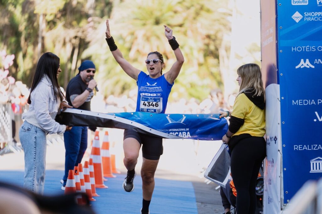 A imagem mostra uma corredora cruzando a linha de chegada da Maratona Internacional de Piracicaba. Ela usa uma camiseta azul com o número 4346, óculos escuros e levanta os dois braços em comemoração. Duas mulheres seguram uma faixa azul da maratona, enquanto um homem ao fundo fala ao microfone. O percurso está cercado por cones laranjas e árvores, e há um painel de patrocinadores à direita.