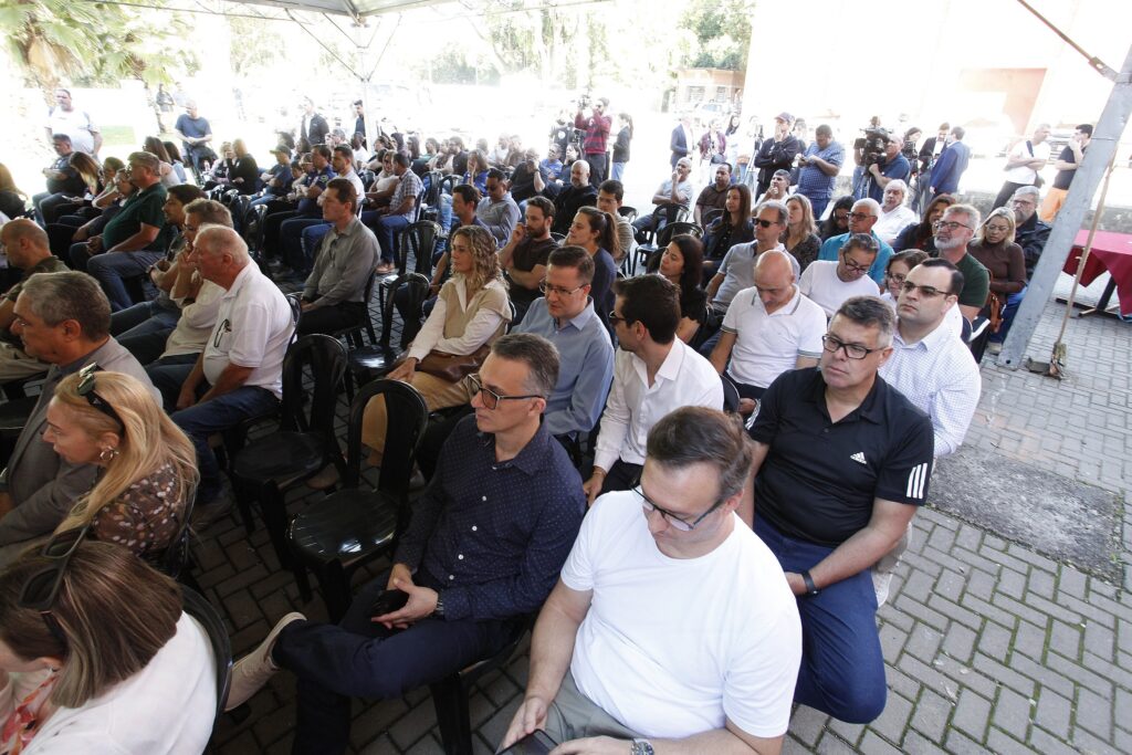 A imagem registra uma plateia atenta durante um evento ao ar livre, possivelmente uma cerimônia ou apresentação oficial, realizada sob uma tenda. O público: Vemos dezenas de pessoas sentadas em cadeiras de plástico pretas, organizadas em fileiras. O ambiente é predominantemente masculino, com vários homens usando camisas sociais, polos e óculos. Há também algumas mulheres presentes, distribuídas entre o público. O clima: As pessoas parecem concentradas, olhando em direção a um orador ou palco que está fora do enquadramento, à frente de todos. O local: O piso é de pavimentos intertravados de concreto. Ao fundo, percebe-se uma área aberta com árvores e iluminação natural intensa, contrastando com a sombra projetada pela tenda onde o público está sentado. Ao lado direito, nota-se uma mesa coberta por uma toalha vermelha, sugerindo a estrutura de apoio ao evento.