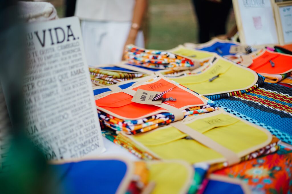 Esta imagem captura uma bancada de feira ao ar livre repleta de artigos têxteis artesanais coloridos, exibindo um letreiro inspirador e uma variedade de acessórios organizados para venda ao público. A composição é baseada em uma profundidade de campo rasa, focando no primeiro plano e desfocando progressivamente o fundo, o que cria um destaque atraente nos produtos. Os itens estão dispostos de maneira organizada em uma mesa coberta, sugerindo uma configuração de mercado ou evento cultural. O letreiro posicionado à esquerda estabelece o contexto temático, enquanto a disposição dos tecidos em camadas e grupos convida o olhar do espectador a percorrer toda a variedade de texturas e padrões presentes na bancada. Os principais objetos são bolsas ou estojos de tecido dobrados, caracterizados por suas cores vivas como laranja, amarelo e azul, frequentemente combinados com padrões estampados que remetem a motivos tradicionais ou artesanais. Alguns itens possuem etiquetas de papel presas por cordões, indicando preços ou marcas, o que reforça a natureza comercial do objeto. O material parece ser um tecido resistente, talvez algodão ou lona, com acabamentos bem definidos que sugerem uma confecção manual cuidadosa e atenciosa aos detalhes. A fotografia utiliza uma técnica de foco seletivo que separa o objeto central do ambiente circundante, uma escolha clássica na fotografia de produtos para conferir um aspecto profissional e artístico. O estilo é documental e naturalista, priorizando a luz ambiente sobre o uso de iluminação artificial pesada. A execução técnica destaca a nitidez das texturas nos tecidos em primeiro plano, permitindo observar a trama do material e as dobras, enquanto a saturação das cores é realçada pela luz suave do dia. O cenário parece ser uma feira de artesanato ou um evento social diurno, onde a luz natural difusa cria uma atmosfera acolhedora e vibrante. A iluminação é suave, possivelmente vinda de uma luz solar filtrada ou de um céu nublado, o que evita sombras duras sobre os produtos e permite que as cores dos tecidos se tornem o elemento dominante da cena. O fundo levemente desfocado sugere a presença de pessoas ou outros elementos de uma praça, contribuindo para uma sensação de movimento e vida coletiva, resultando em um clima alegre, criativo e artesanal.