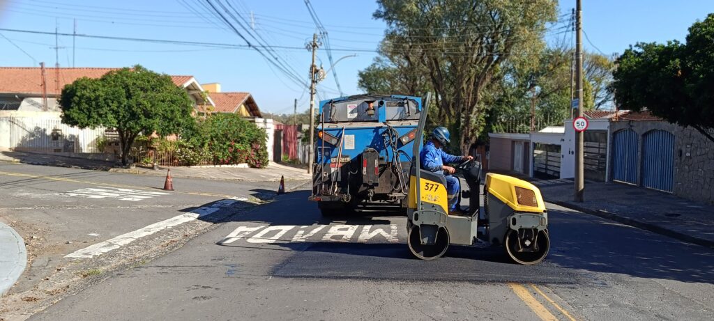 vemos um trabalhador com uniforme azul e capacete operando um pequeno rolo compressor amarelo, identificado pelo número “377”, para compactar o asfalto recém-aplicado em uma rua de bairro. À frente da máquina, há um caminhão azul de asfalto com equipamentos acoplados na traseira, enquanto cones de trânsito delimitam a área de serviço. O pavimento exibe a palavra “PARE” pintada e uma placa de limite de velocidade de 50 km/h aparece ao lado, compondo o cenário urbano com casas de telhado cerâmico e árvores ao redor.