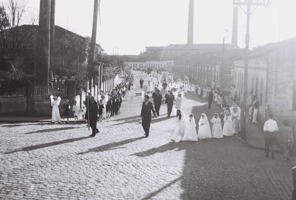 A fotografia mostra uma procissão religiosa antiga em uma rua de paralelepípedos, registrada em preto e branco. O grupo é formado por crianças vestidas de branco, provavelmente em trajes de primeira comunhão, acompanhadas por adultos de terno escuro. Ao fundo, vê-se uma fileira de casas simples e chaminés industriais, sugerindo que o evento ocorre em uma cidade operária.