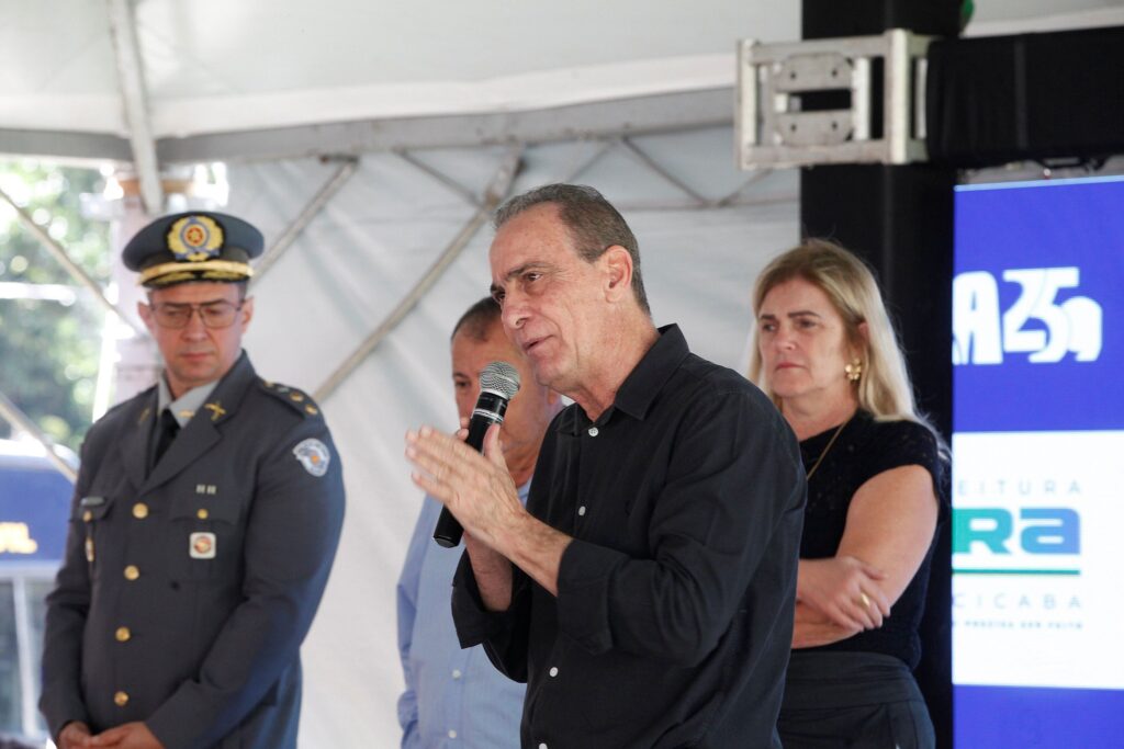 A foto mostra um momento de um evento oficial ou pronunciamento. No centro, um homem de camisa social preta está falando ao microfone, com um gesto das mãos que sugere que ele está explicando ou enfatizando algo. Ao lado dele, estão outras pessoas compondo a cena: À esquerda: Um policial militar uniformizado, em posição de respeito, e parte de outro homem vestindo uma camisa azul clara. À direita: Uma mulher de cabelos loiros e blusa preta, que parece estar ouvindo atentamente. Ao fundo, há um painel digital azul com uma parte da marca "Pira" (provavelmente uma referência à Prefeitura de Piracicaba, como notado anteriormente), indicando que se trata de uma cerimônia pública ou institucional.