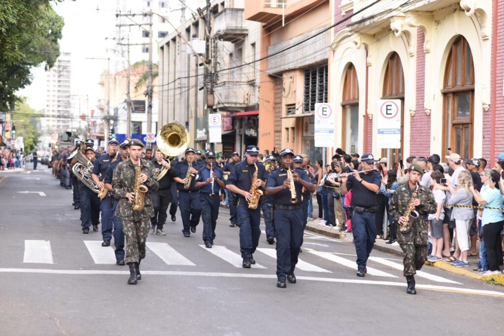 A imagem mostra uma banda marcial desfilando por uma rua movimentada. Os músicos usam uniformes azul-marinho e tocam saxofones, clarinetes, flautas e instrumentos de metal, incluindo uma grande tuba. À frente e nas laterais, há integrantes usando uniformes camuflados e tocando saxofone. O público acompanha o desfile nas calçadas, separado por uma fita de segurança, enquanto prédios históricos, placas de estacionamento e muitos fios elétricos compõem o cenário urbano.