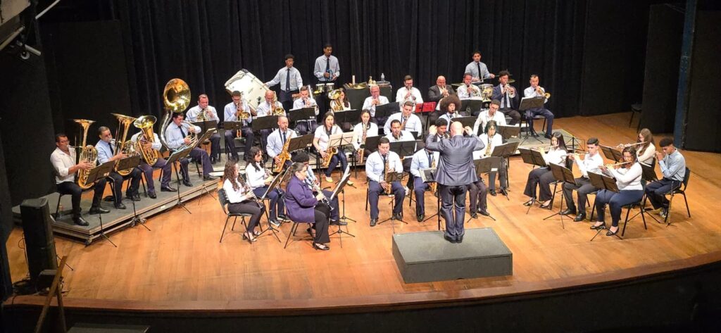 A imagem mostra uma grande banda ou orquestra de sopros se apresentando em um palco de teatro. Os músicos estão sentados em várias fileiras, tocando instrumentos como trompetes, trombones, saxofones, tubas, eufônios e percussão. Todos têm partituras em estantes à frente. No centro do palco, um maestro de terno está de costas para o público, conduzindo o grupo com os braços erguidos. O palco tem piso de madeira, cortinas pretas ao fundo e iluminação quente.