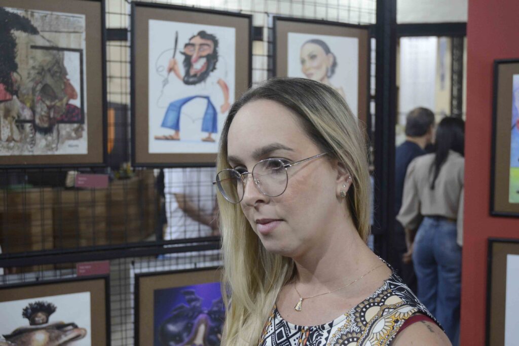 A imagem mostra uma mulher loira usando óculos grandes e uma blusa estampada, em primeiro plano, no lado direito. Ela parece estar sorrindo ou falando. Ao fundo, há uma parede com vários pôsteres e fotografias de pessoas, além de prateleiras com objetos coloridos. A cena parece acontecer em um ambiente interno, possivelmente uma loja, estúdio ou espaço cultural.