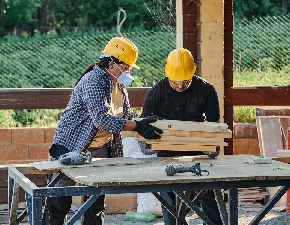 A imagem mostra dois trabalhadores em uma obra ou oficina de marcenaria. Ambos usam capacetes amarelos; a pessoa à esquerda também veste óculos de proteção, máscara, luvas e camisa xadrez. Eles estão manuseando uma estrutura ou peças de madeira sobre uma bancada. Ao redor, há ferramentas elétricas, tábuas e materiais de construção. Ao fundo, vê-se uma cerca verde e bastante vegetação.