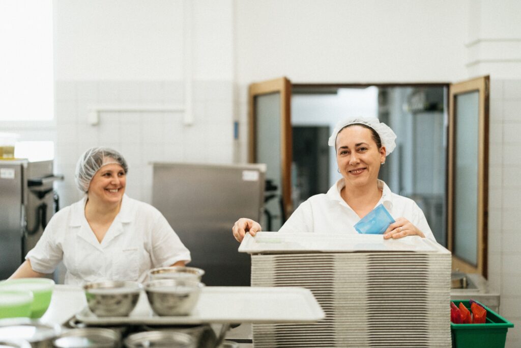 A imagem mostra duas mulheres trabalhando em uma cozinha profissional. Ambas vestem uniformes brancos e toucas de proteção, típicos de ambientes de preparo de alimentos. Uma delas está sorrindo, enquanto a outra segura uma pilha de bandejas e um item azul, possivelmente um pano ou embalagem. O ambiente é limpo e bem iluminado, com utensílios de aço inox sobre a bancada — transmitindo uma atmosfera de organização e higiene.