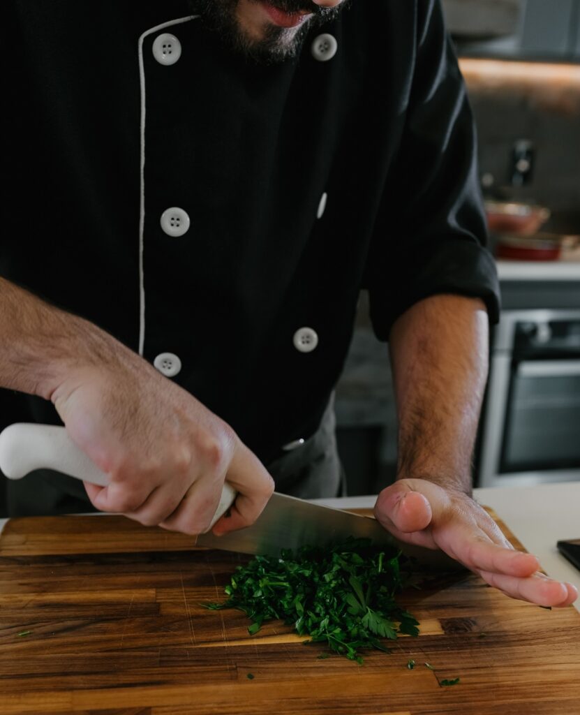 Esta imagem captura uma cena clássica de cozinha profissional ou caseira, focada na técnica de preparo. Aqui estão os detalhes: A Ação: Um chef está picando ervas frescas (parecem salsinha ou coentro) sobre uma tábua de madeira. A mão direita segura firmemente o cabo da faca, enquanto a mão esquerda, em posição de "garra" (uma técnica fundamental para segurança na cozinha), estabiliza o maço de ervas. O Uniforme: A pessoa veste uma dólmã (jaqueta de chef) preta, que possui botões brancos e um detalhe de linha branca na gola, conferindo um visual elegante e profissional. Ambiente: O foco está na bancada de trabalho. Ao fundo, levemente desfocado, é possível ver um ambiente de cozinha moderna com um forno ou fogão embutido, sugerindo que o trabalho está acontecendo em um espaço funcional e bem equipado. Sensação: A foto transmite precisão e cuidado, destacando a habilidade manual necessária na culinária.