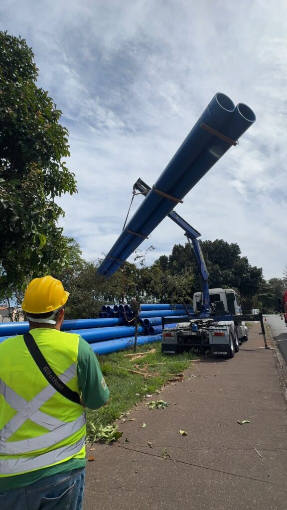 A imagem mostra uma cena de trabalho ao ar livre, provavelmente em uma área urbana ou suburbana, com foco na movimentação de tubos grandes e azuis. A perspectiva é do ponto de vista de alguém que está observando a operação. No primeiro plano, vemos a parte traseira de um homem vestindo um colete de segurança de alta visibilidade na cor verde limão com detalhes em amarelo e branco refletivos. Ele usa um capacete de segurança amarelo e tem a palavra "Alfatronic" escrita em sua faixa refletiva. Sua mão direita está ligeiramente visível, e ele parece estar observando a ação. O corpo principal da imagem é dominado por tubos azuis. Uma fileira de tubos azuis está empilhada no chão, à esquerda e ao fundo. Acima desta pilha, um guindaste, preso a um caminhão branco da Scania com a marca da empresa "GUEVARA" visível na traseira, está erguendo vários tubos azuis grandes e longos no ar. Dois desses tubos estão bem evidentes, suspensos horizontalmente por cabos e suportes de madeira presos ao guindaste. O céu está visível acima dos tubos, com nuvens brancas dispersas sobre um fundo azul claro. A cena também inclui vegetação, com árvores frondosas de folhas verdes escuras e algumas folhas caídas no chão, sugerindo um ambiente natural. Há uma calçada de concreto ou asfalto cinza à direita, que margeia a área de trabalho e o gramado onde os tubos estão. Uma estrada asfaltada é visível ao fundo à direita, com um reflexo do céu ou de outra estrutura. A iluminação geral sugere um dia claro, possivelmente com sol em alguma parte do céu, mas com nuvens que suavizam a luz direta. A composição foca na atividade de carregamento e na magnitude dos tubos, com o trabalhador servindo como ponto de referência humano para a escala da operação. A cor azul vibrante dos tubos contrasta com os tons naturais do verde e do céu, além do branco do caminhão e dos elementos refletivos do colete.