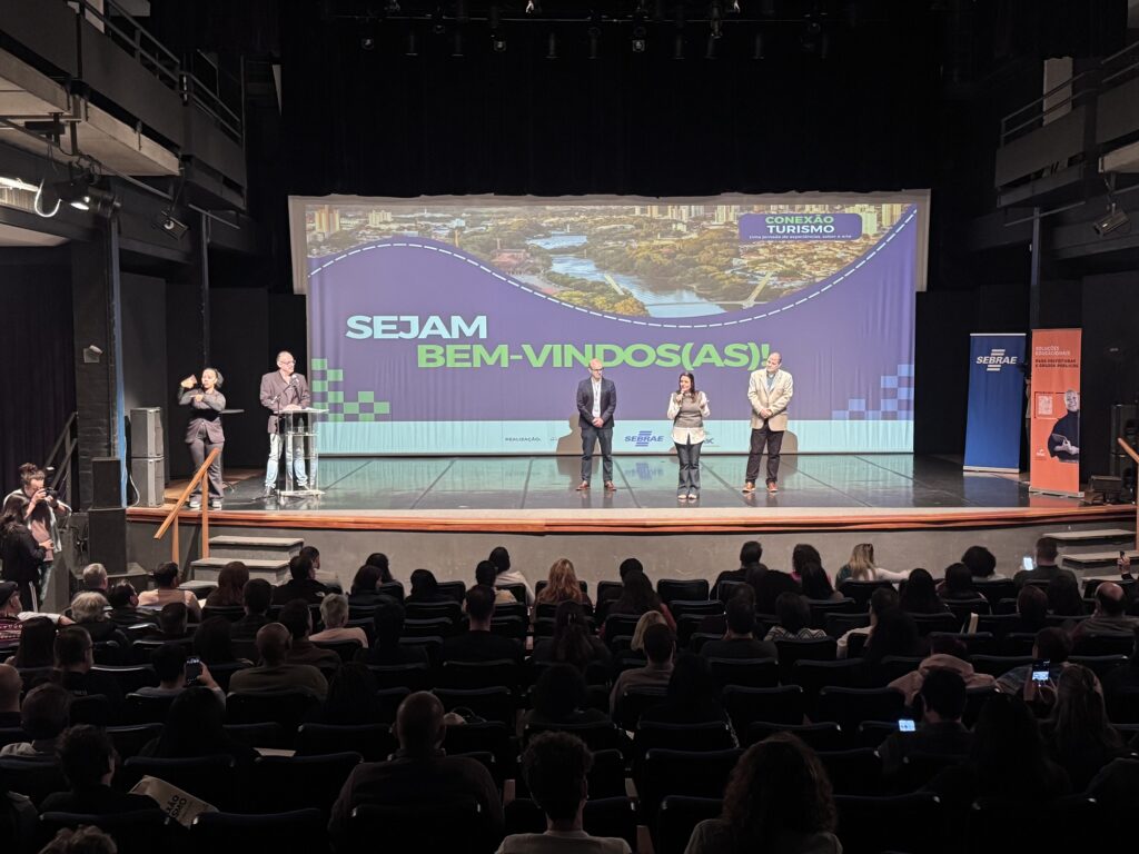 A imagem mostra um evento institucional realizado em um teatro ou auditório, com um público numeroso ocupando as poltronas e acompanhando as atividades no palco.
No palco, há quatro pessoas em destaque, aparentemente participando da abertura ou apresentação do evento. À esquerda, uma pessoa faz uma apresentação em língua de sinais, enquanto outra está junto a um púlpito. Ao centro e à direita, outras duas pessoas estão posicionadas diante do público.
Ao fundo, um grande telão exibe a mensagem “SEJAM BEM-VINDOS(AS!)” e a identidade visual do evento “Conexão Turismo — Uma jornada de experiências, sabor e arte”, com uma imagem aérea de uma área urbana e paisagens naturais. Também aparecem as marcas do Sebrae e referências ao Senac.
Nas laterais do palco, há banners institucionais, incluindo um do Sebrae. Algumas pessoas na plateia seguram celulares, registrando o momento.
A cena transmite um clima de evento profissional, receptivo e voltado ao turismo, capacitação e integração de participantes.