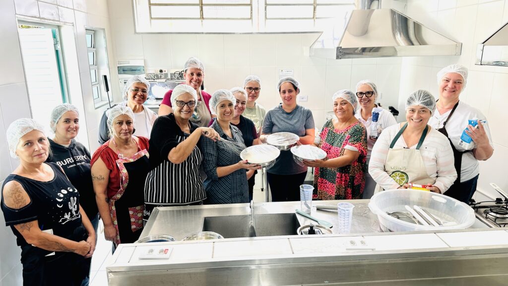 A imagem mostra um grupo de treze pessoas reunidas em uma cozinha ampla e bem equipada, todas usando toucas higiênicas e algumas vestindo aventais, transmitindo a ideia de participação em uma oficina ou aula de culinária; alguns seguram tigelas cobertas com plástico filme, outros utensílios como batedores e recipientes, reforçando o clima de colaboração e aprendizado gastronômico em um ambiente profissional e organizado.