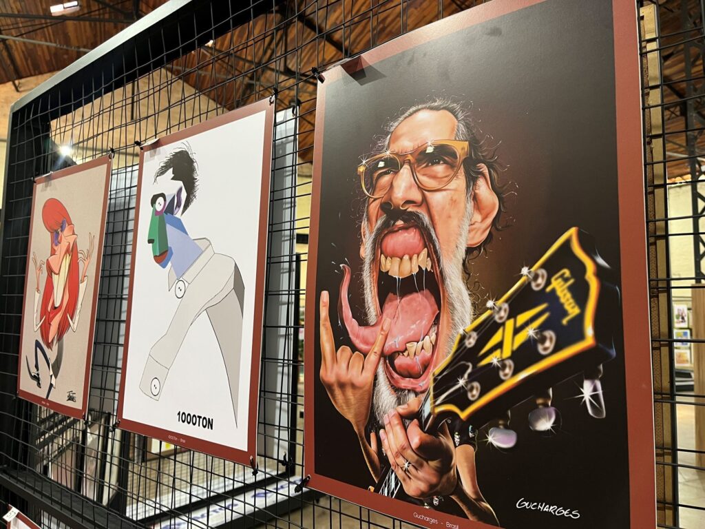 A imagem mostra uma exposição de ilustrações e caricaturas penduradas em uma estrutura metálica preta, dentro de um espaço com teto de madeira. Em destaque, há um retrato caricatural de um homem de cabelos grisalhos, usando óculos grandes e fazendo uma expressão exagerada, com a boca aberta e a língua para fora. Ele segura um instrumento musical escuro com detalhes amarelos. Na parte inferior da obra, aparece a assinatura “Gucharges”. Ao lado, há uma ilustração minimalista de uma figura com cabeça geométrica e o texto “1000TON”. Mais à esquerda, vê-se outra obra colorida com uma personagem de cabelos vermelhos.