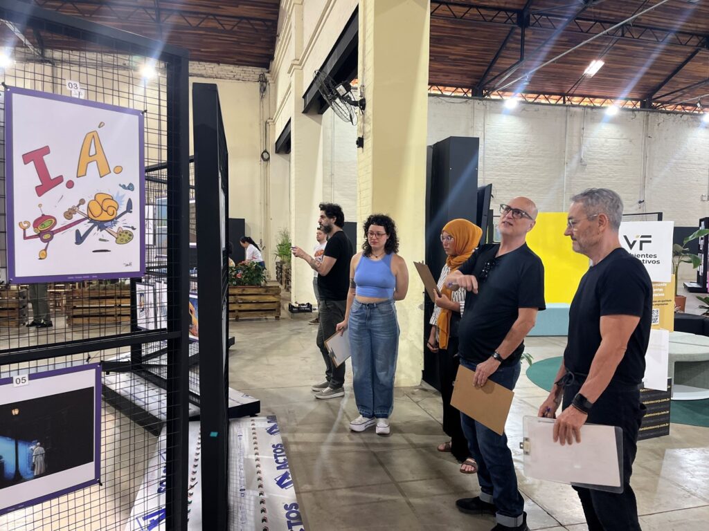 A imagem mostra um grupo de pessoas visitando uma exposição em um amplo galpão com paredes claras, teto de madeira e estrutura metálica. À esquerda, há painéis com obras de arte, incluindo uma ilustração colorida com as letras “I.A.” e outra imagem em preto e branco. No centro e à direita, visitantes observam os trabalhos; alguns seguram pranchetas, sugerindo uma avaliação ou visita guiada. Entre eles estão uma mulher de blusa azul, uma mulher usando lenço mostarda e dois homens vestidos de preto. Ao fundo, aparecem outros estandes, plantas, caixas e um painel amarelo com informações sobre uma empresa.