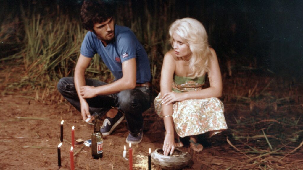 A imagem mostra duas pessoas sentadas no chão de uma área rural, cercadas por vegetação alta. Um homem de camiseta azul e calça escura olha para uma mulher loira, que usa um vestido estampado com faixa verde. Ao redor delas há várias velas acesas, uma garrafa e alguns objetos no chão, criando uma atmosfera misteriosa, como se participassem de um ritual ou cena de filme.