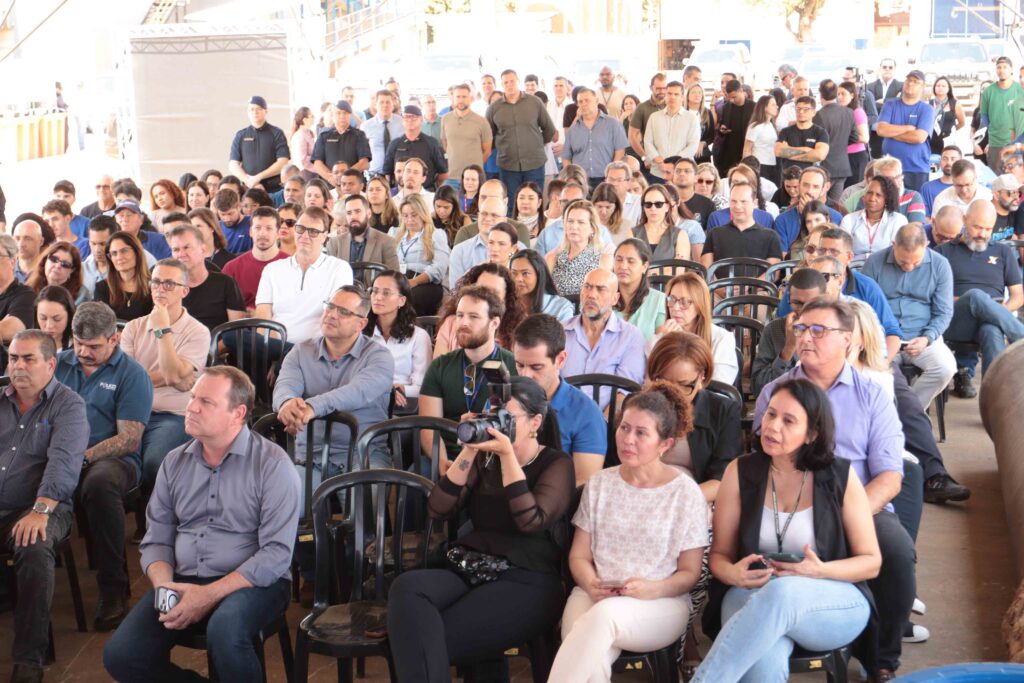 A imagem mostra um grande grupo de pessoas reunido em um auditório ou salão amplo, provavelmente durante um evento institucional ou uma reunião. Muitas pessoas estão sentadas em cadeiras, enquanto outras permanecem em pé ao fundo. Várias usam crachás e camisetas ou uniformes com logotipos, sugerindo que participam da mesma organização. O ambiente é bem iluminado, com janelas e estruturas metálicas ao fundo. A fotografia está superexposta em algumas áreas e registra uma plateia diversificada, formada principalmente por adultos.