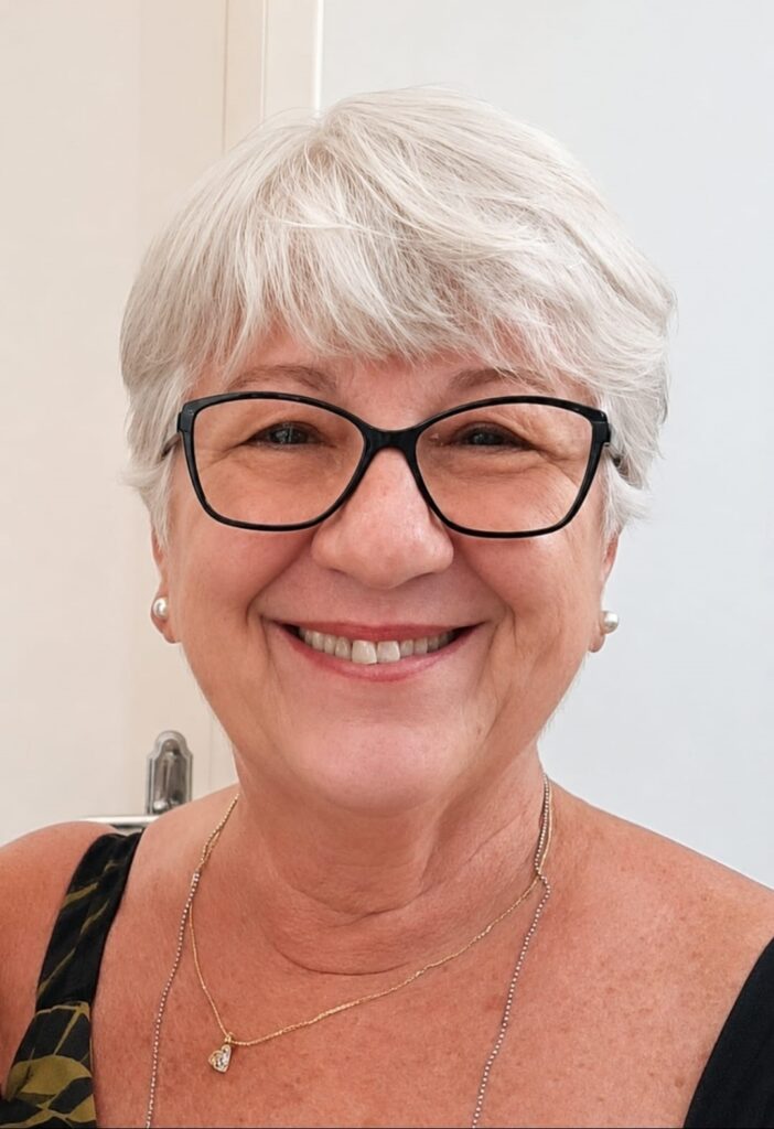 Na foto há o rosto de uma mulher sorridente, enquadramento próximo (close-up): Cabelo curto e grisalho, penteado com franja suave. Usa óculos de aro preto com lente transparente. Sorriso aberto, dentes visíveis; expressão calorosa e amigável. Brincos de pérola pequenos e colares delicados (um com pingente de coração). Pele com sinais naturais de envelhecimento (linhas de expressão ao redor dos olhos e da boca). Roupas: alça aparente de uma peça sem mangas com estampa escura. Fundo claro e neutro, iluminação suave que destaca o rosto.