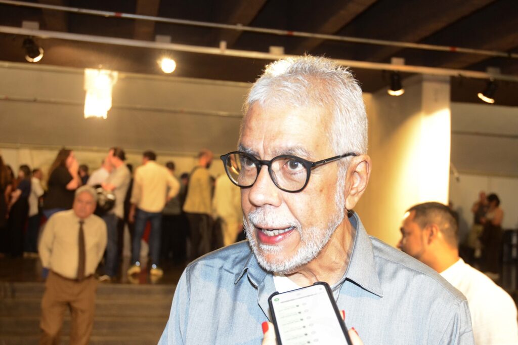 A imagem mostra, em primeiro plano, um homem idoso de cabelos e barba brancos, usando óculos de armação preta e camisa azul-clara. Ele parece estar sendo entrevistado, pois há um celular com a tela voltada para ele, segurado por outra pessoa. O ambiente é um espaço amplo, com teto de madeira, iluminação por refletores e várias pessoas conversando ao fundo. A fotografia tem iluminação forte e quente, típica de um evento ou exposição.