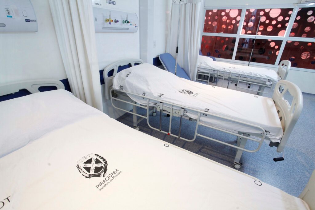 Esta imagem retrata um quarto de enfermaria hospitalar limpo e organizado, apresentando várias camas de hospital dispostas lado a lado, prontas para receber pacientes, dentro de uma instalação médica funcional e bem cuidada. A composição da cena é estruturada em torno de três leitos hospitalares posicionados de forma paralela, ocupando o centro e as laterais do ambiente. A perspectiva é capturada de um ângulo levemente elevado, o que permite uma visualização clara da disposição das camas e do espaço entre elas. Cortinas brancas suspensas servem como divisórias de privacidade, criando delimitações individuais para cada leito. O uso de linhas diagonais criadas pela posição das camas confere uma sensação de profundidade e ordem ao espaço, guiando o olhar do espectador desde a cama mais próxima em primeiro plano até a área da janela no fundo. As camas hospitalares são o elemento central, possuindo estruturas metálicas modernas com rodas e grades laterais ajustáveis. Elas estão cobertas com lençóis brancos impecáveis que apresentam o brasão da prefeitura de Piracicaba impresso de forma nítida. O material das roupas de cama parece ser algodão padrão hospitalar, transmitindo uma imagem de higiene rigorosa. Cada cama está equipada com mecanismos para ajuste de altura e encosto, denotando sua funcionalidade clínica. Não há pessoas ou objetos pessoais visíveis, conferindo ao ambiente um aspecto estéril e de espera. A imagem é uma fotografia digital de alta definição, caracterizada por uma técnica clara que foca na representação realista do ambiente. A profundidade de campo é mantida de modo que tanto as camas em primeiro plano quanto os detalhes estruturais do fundo permanecem nítidos. As texturas, como o metal frio das estruturas das camas e a suavidade dos tecidos, são reproduzidas com fidelidade. O estilo é puramente funcional e documental, enfatizando a organização, a limpeza e a preparação do espaço hospitalar sem artifícios artísticos excessivos. O ambiente é um quarto de hospital contemporâneo, iluminado por uma luz difusa e clara, provavelmente proveniente de lâmpadas fluorescentes de teto, que garante boa visibilidade em todo o recinto. As paredes são de um branco clínico, contribuindo para a atmosfera de assepsia. Ao fundo, uma grande janela com uma película adesiva decorativa em tons avermelhados com padrões circulares oferece um contraste visual, suavizando a luz externa e trazendo um elemento de design singular à arquitetura. O piso é de material liso e claro, condizente com as normas sanitárias. O clima é de serenidade, ordem e prontidão, transmitindo uma sensação de um espaço preparado profissionalmente para o cuidado de pacientes.