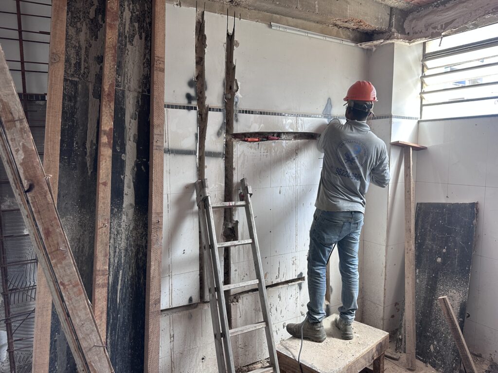 Na cena, um trabalhador da construção civil veste capacete vermelho, camisa cinza de mangas compridas, jeans e botas, enquanto atua sobre uma plataforma de madeira em frente a uma parede revestida com azulejos brancos e uma faixa decorativa. Ele parece cortar ou ajustar a superfície, em meio a vigas de madeira expostas, materiais espalhados e uma escada metálica apoiada na parede. A iluminação natural entra por uma janela com grades horizontais, destacando o ambiente em reforma e transmitindo a atmosfera de esforço manual e transformação do espaço.