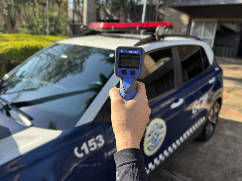 A imagem mostra uma pessoa sentada em um veículo, segurando um oxímetro de pulso azul no dedo. O aparelho exibe a palavra “Inoximeters” e parece estar em uma tela de teste. Ao fundo, há uma área externa desfocada, com árvores, gramado e um caminho ou rua.