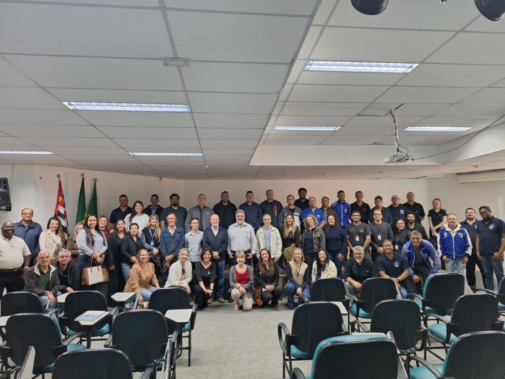 Uma grande fotografia de grupo registra um momento profissional em uma sala de conferências ampla e iluminada, reunindo dezenas de homens e mulheres sorridentes, alinhados para uma imagem coletiva memorável. A composição da cena é organizada de forma estruturada, com as pessoas dispostas em várias fileiras, ocupando toda a largura do ambiente. O grupo principal está de pé, enquanto algumas pessoas se encontram na primeira fila, em pé ou sentadas, criando uma distribuição equilibrada e coesa. O enquadramento é amplo, capturando o espaço interno completo, incluindo cadeiras dispostas em fileiras no primeiro plano, o que conduz o olhar do observador diretamente para o centro do grupo centralizado ao fundo da sala. Os sujeitos da imagem são um grupo diversificado de adultos, composto por homens e mulheres de várias idades, apresentando uma mistura de estilos de vestimenta que variam do casual ao casual de negócios. Muitos usam camisas de botão, jaquetas, suéteres e roupas sociais, refletindo um ambiente de trabalho ou reunião de conferência. As expressões são predominantemente amigáveis, com sorrisos e olhares voltados para a câmera, denotando uma atmosfera de cooperação e harmonia entre os participantes. Esta é uma fotografia digital capturada com clareza, exibindo técnicas de fotografia documental padrão. O foco é nítido em todos os planos, garantindo que os rostos e detalhes do grupo sejam visíveis. A execução privilegia a iluminação uniforme vinda dos painéis de luz do teto, o que reduz sombras duras e mantém o tom natural e profissional da imagem, condizente com registros institucionais ou corporativos. O cenário é uma sala de reuniões ou auditório funcional, com paredes de tons claros, teto de placas e um carpete cinza. A presença de duas bandeiras, uma delas sendo a bandeira do estado de São Paulo, posicionadas à esquerda, sugere um contexto institucional ou governamental. A iluminação artificial é brilhante e onipresente, resultando em uma atmosfera clara, organizada e séria, típica de um espaço de trabalho destinado a encontros, treinamentos ou eventos coletivos de um grupo de trabalho.