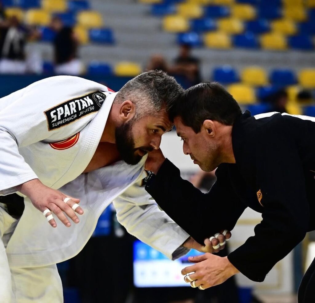 Dois lutadores de jiu-jitsu em um momento de intensa concentração se encaram durante uma competição.

A imagem captura um close-up de dois homens competindo em uma luta de jiu-jitsu, com seus rostos muito próximos, transmitindo uma sensação de tensão e foco intenso. O lutador da esquerda veste um quimono branco, com etiquetas visíveis que dizem "SPARTAN" e "SUPREMACIA JIU-JITSU". Ele tem barba grisalha e cabelos curtos e grisalhos, com os dedos da mão esquerda enfaixados em branco. O lutador da direita veste um quimono preto e tem os cabelos escuros e curtos, com os dedos da mão direita também enfaixados em branco, enquanto segura o braço do oponente. Ambos parecem estar em uma posição de agarrar ou de início de movimento, o que sugere que a luta está em andamento. O fundo está desfocado, mas exibe fileiras de assentos em tons de azul e amarelo, indicando que estão em uma arena ou ginásio. A iluminação foca nos lutadores, destacando a expressão concentrada em seus rostos e a textura dos seus quimonos. A atmosfera é de seriedade e competitividade, com um humor centrado na intensidade do confronto esportivo.