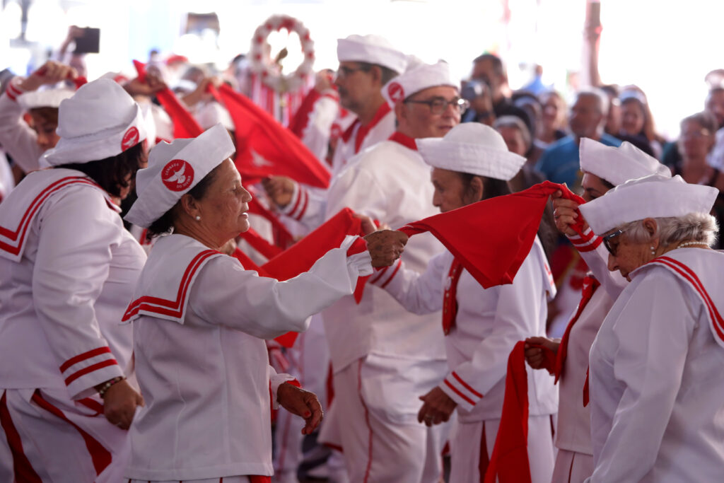 Na imagem, um grupo de pessoas vestindo uniformes brancos com detalhes vermelhos e chapéus brancos participa de um evento. O foco principal está em várias mulheres idosas que seguram bandeiras ou lenços vermelhos vibrantes. Seus uniformes consistem em jaquetas brancas com golas e punhos vermelhos, e possivelmente saias ou calças brancas com listras vermelhas verticais. Os chapéus são brancos, quadrados ou retangulares, com um emblema que parece ser um pássaro branco dentro de um círculo vermelho. As cores predominantes são o branco e o vermelho, criando um contraste forte e festivo. O fundo é desfocado, sugerindo um ambiente externo ou um local com grande concentração de pessoas, possivelmente uma celebração ou procissão. A iluminação é clara, provavelmente luz natural, destacando os rostos das mulheres e a vivacidade das bandeiras vermelhas. A composição coloca as mulheres em primeiro plano, muitas delas interagindo umas com as outras ou com os lenços vermelhos, sugerindo movimento e um ritual. A mulher mais à esquerda na imagem, em destaque, está virada para o lado, segurando um lenço vermelho com ambas as mãos e olhando para frente. Ao seu lado, outra mulher idosa, com óculos escuros e um colar de pérolas, também está envolvida na manipulação de um lenço vermelho. A profundidade de campo é rasa, mantendo o foco nas figuras centrais e desfocando o restante do grupo e o fundo. A textura dos tecidos dos uniformes e das bandeiras é visível, assim como as rugas e a pele das mulheres, conferindo um realismo à cena. O clima geral da imagem parece ser de alegria, comunidade e participação em uma tradição.