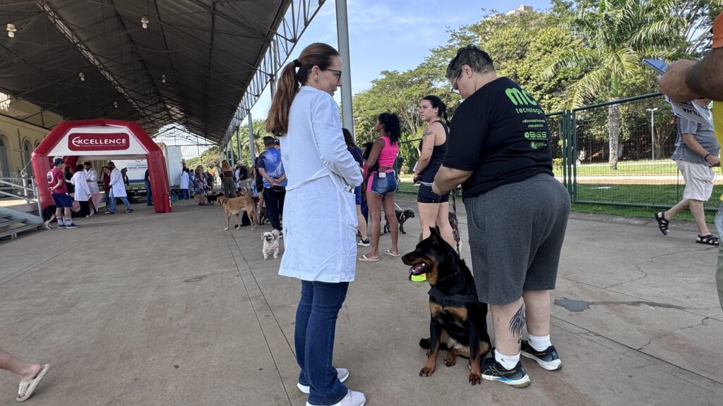Na imagem, vemos um evento ao ar livre realizado sob uma grande cobertura, reunindo várias pessoas acompanhadas de seus cães. Alguns participantes estão em fila, enquanto outros vestem jalecos brancos, sugerindo a presença de profissionais veterinários ou organizadores. Um arco inflável vermelho com a palavra “EXCELLENCE” marca a entrada ou área de registro. O ambiente é amplo e iluminado, com árvores e gramado ao fundo, transmitindo a ideia de um espaço comunitário.