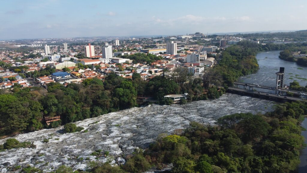 A imagem apresenta uma vista aérea panorâmica de uma cidade brasileira, com destaque para um amplo curso d’água e uma área de corredeiras ou queda d’água em primeiro plano.
Na parte central e ao fundo, observa-se uma paisagem urbana bastante desenvolvida, com casas de diferentes padrões, edifícios residenciais e comerciais, áreas industriais e ruas arborizadas. A cidade se estende até o horizonte, mostrando sua dimensão e ocupação territorial.
Em primeiro plano, a vegetação acompanha as margens do rio, criando um contraste marcante entre a natureza e a área urbana. O rio apresenta grande volume de água e corredeiras, enquanto uma ponte atravessa o curso d’água à direita da imagem.
O enquadramento aéreo permite perceber, de forma bastante clara, a relação entre cidade, rio, áreas verdes e infraestrutura urbana. O céu aberto e a iluminação natural também contribuem para uma sensação de amplitude.
Pelo aspecto da paisagem, a imagem é particularmente adequada para representar turismo urbano, patrimônio natural e a relação histórica de uma cidade com seu rio.