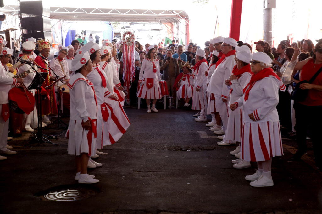O evento retratado na imagem parece ser uma celebração ou procissão, com várias pessoas vestindo uniformes brancos e vermelhos. A cena se passa ao ar livre, com estruturas temporárias de cobertura e um fundo que sugere uma área urbana ou um local de evento. A iluminação indica que é dia, possivelmente com luz solar forte que cria algumas sombras. Os participantes principais estão divididos em dois grupos, formando um corredor central. Muitos estão usando trajes brancos com detalhes em vermelho, como lenços, faixas, punhos de manga e bordas de saia ou calça. As mulheres usam saias plissadas com listras verticais vermelhas e brancas, e algumas usam chapéus brancos com um emblema vermelho circular. Os homens, em sua maioria, usam calças brancas, camisas brancas e chapéus brancos semelhantes. Há uma figura central, possivelmente um líder ou figura de honra, usando uma vestimenta vermelha e dourada elaborada, com uma coroa. Na lateral esquerda, observa-se um grupo de músicos com tambores vermelhos e instrumentos musicais, como um violão. Eles também parecem estar vestindo uniformes semelhantes. Na lateral direita, há uma multidão de espectadores, observando o evento. As cores predominantes são o branco e o vermelho dos uniformes, contrastando com o asfalto escuro do chão e as sombras. A composição da imagem é simétrica, com os dois grupos de participantes alinhados um em frente ao outro, guiando o olhar do espectador para o fundo. A perspectiva é ligeiramente baixa, dando ênfase às figuras em primeiro plano e à grandiosidade do evento. Um detalhe interessante no chão em primeiro plano é uma tampa de bueiro metálica com um padrão circular, que reflete a luz. A atmosfera geral parece ser festiva e comunitária.