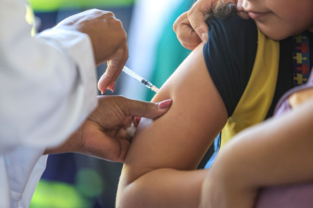 Uma fotografia capturada em um ângulo próximo mostra o momento preciso em que uma profissional de saúde administra uma vacina no braço de uma criança, destacando o cuidado e a assistência médica. A composição é focada na ação da vacinação, com o enquadramento fechado que privilegia a interação entre as mãos da profissional e o braço da criança. A profissional de saúde está à esquerda, com suas mãos posicionadas de forma técnica e cuidadosa para realizar o procedimento, enquanto o braço da criança ocupa o centro e o lado direito do quadro. A disposição dos elementos enfatiza a precisão do movimento, com a agulha sendo inserida suavemente na pele, criando uma narrativa visual de saúde pública e prevenção. Os sujeitos principais são a profissional de saúde e a criança. A profissional, vestida com um jaleco branco, demonstra firmeza e delicadeza em suas mãos, que possuem unhas pintadas de forma discreta. A criança, que aparece parcialmente, usa uma camiseta azul e um colete amarelo com estampas de peças de quebra-cabeça, símbolo associado à conscientização sobre o autismo. Seu rosto, visto apenas em parte na lateral superior, transmite uma postura calma, enquanto seu braço permanece relaxado para receber a dose, indicando um momento de cooperação entre o paciente e o profissional. Esta imagem é uma fotografia documental que utiliza uma profundidade de campo rasa, técnica comum em registros fotográficos para isolar o objeto de interesse. O foco está nitidamente concentrado no ponto de contato entre a agulha e o braço, deixando o fundo e outras partes do cenário com um leve desfoque, o que confere um aspecto mais profissional e humanizado à cena. As cores são naturais, com o branco do jaleco servindo como ponto de contraste para os tons de pele e as vestimentas coloridas, resultando em uma estética limpa, direta e informativa. O cenário parece ser um ambiente clínico ou de um posto de vacinação, caracterizado por uma iluminação suave e difusa que evita sombras fortes, transmitindo uma sensação de higiene e serenidade. A luz incide de forma uniforme, permitindo que todos os detalhes da ação, como o ângulo da seringa e a posição dos dedos, sejam claramente visíveis. O ambiente sugere um momento cotidiano de atendimento médico, onde a atmosfera é pautada pelo profissionalismo, segurança e responsabilidade. Não há distrações visuais, permitindo que a atenção do observador se concentre totalmente no gesto protetor da vacinação.
