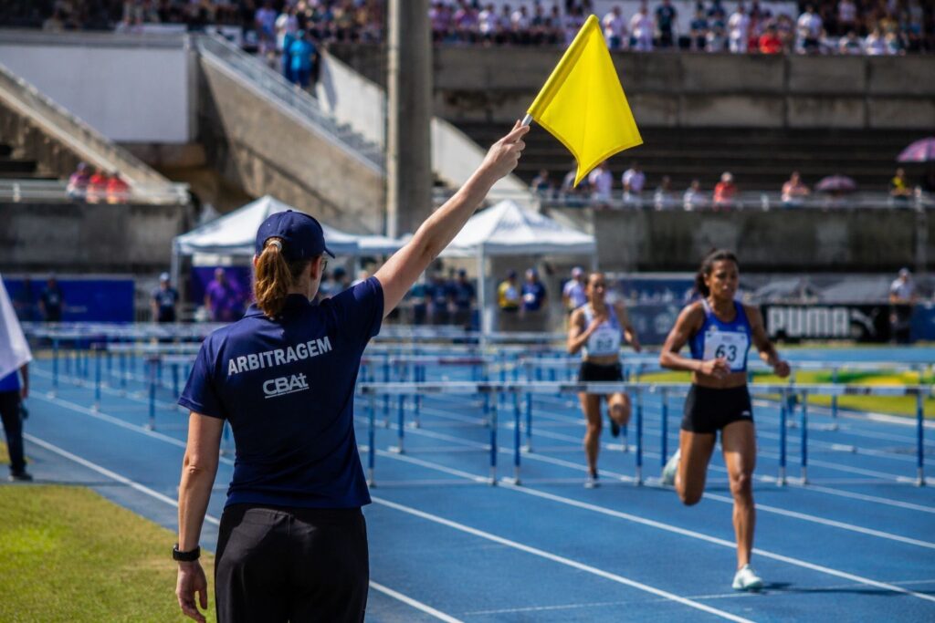 A imagem mostra uma árbitra de atletismo em primeiro plano, vista de costas, levantando uma bandeira amarela. Ela usa boné e camiseta azul-marinho com a palavra “ARBITRAGEM” estampada nas costas. Ao fundo, duas atletas correm em uma pista azul com barreiras. Uma delas usa o número 63 no uniforme. O evento acontece em um estádio ao ar livre, com arquibancadas cheias de espectadores, tendas e estruturas esportivas.