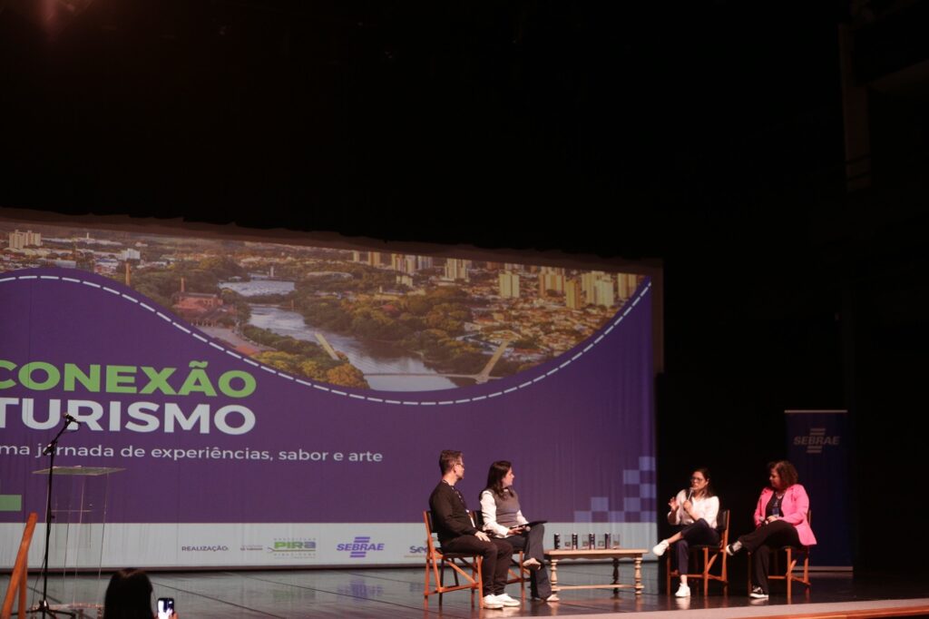 Na imagem vemos um painel de palco de um evento (fundo roxo com uma foto aérea de cidade) com o texto grande “CONEXÃO TURISMO” e logotipos de apoiadores (entre eles SEBRAE). Em primeiro plano, há um encontro em formato de painel: Quatro pessoas sentadas em cadeiras tipo diretor, em linha baixa no palco — à esquerda um homem de roupa escura e ao lado uma mulher com colete cinza; à direita duas mulheres, uma de branco falando ao microfone e outra de rosa ouvindo. A mulher de branco está gesticulando e parece estar respondendo ou explicando algo; as demais escutam atentamente. À esquerda do palco há um pedestal transparente com microfone em pé; ao centro-baixo da imagem nota-se um participante do público registrando a cena com o celular. O ambiente é um auditório escuro, com iluminação focada no palco; ao fundo, além do grande painel, há cortinas pretas cobrindo o teto do palco. A cena transmite uma conversa formal sobre turismo com tom de palestra/painel.