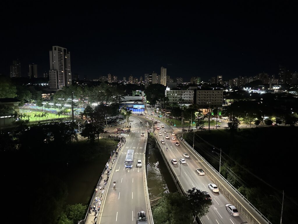 A imagem mostra uma vista panorâmica noturna de uma cidade. Ao fundo, há vários prédios residenciais iluminados contra o céu escuro. Em primeiro plano, uma avenida movimentada atravessa a cena, com carros, ônibus, semáforos e faixas de pedestres. A via passa sobre um canal ou rio estreito, acompanhado por outra pista elevada. À esquerda, há um complexo esportivo com campos iluminados e pessoas praticando atividades. Árvores e áreas verdes aparecem entre as construções e as ruas.
