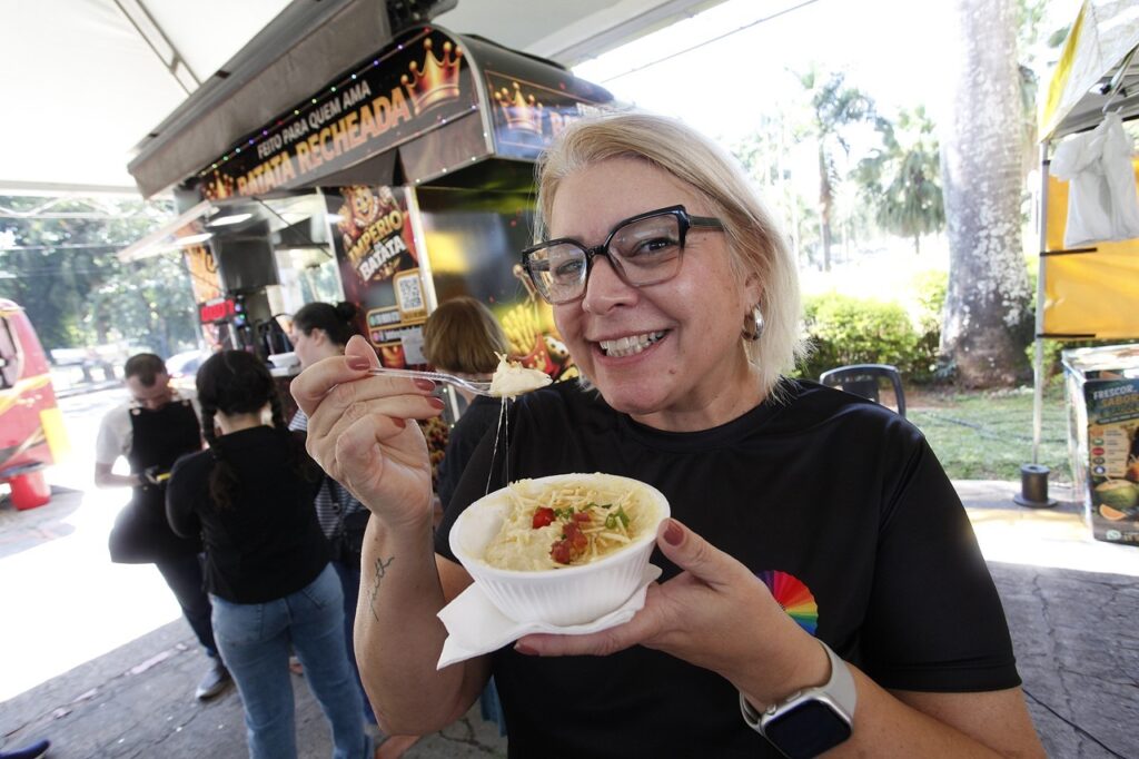 A imagem mostra uma mulher sorridente em primeiro plano, segurando uma tigela de batata recheada (ou purê) com uma colher de plástico. Na colher, é possível ver um pouco do recheio sendo levantado com um fio de queijo derretido. Ela usa óculos, uma blusa preta com um pequeno detalhe colorido na altura do peito, e um relógio tipo smartwatch no pulso. Ao fundo, vemos um food truck de cor escura com detalhes dourados e uma placa escrito "Império da Batata" e "Feito para quem ama batata recheada". O ambiente parece ser um espaço aberto com luz natural, onde outras pessoas estão presentes, incluindo alguém de costas, vestindo preto, que parece ser um atendente ou cliente próximo ao trailer. O cenário sugere um evento de alimentação ao ar livre.