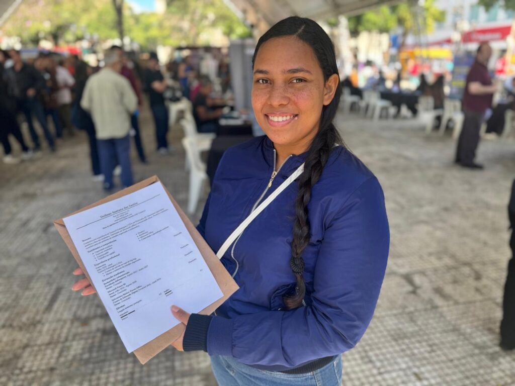 A imagem mostra uma jovem sorrindo, em primeiro plano, segurando uma pasta com um currículo impresso. Ela veste uma jaqueta azul-marinho e está em um espaço aberto, com várias pessoas ao fundo, mesas, cadeiras e barracas, em um ambiente movimentado que aparenta ser uma feira ou evento de oportunidades.