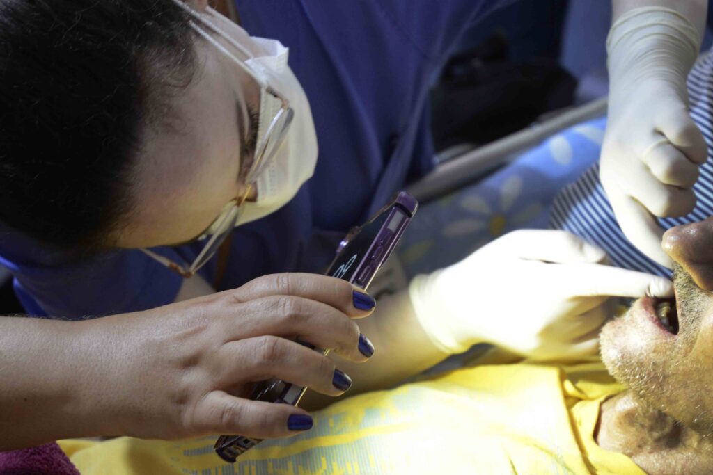 A imagem mostra um atendimento de vacinação. Uma profissional de saúde, usando luvas e avental, aplica uma injeção no braço de uma pessoa sentada. A pessoa veste uma camiseta azul e segura um cartão ou documento de vacinação. Ao fundo, há uma parede azul e uma mesa com materiais médicos. A cena está registrada em close, destacando o procedimento e os braços das duas pessoas.