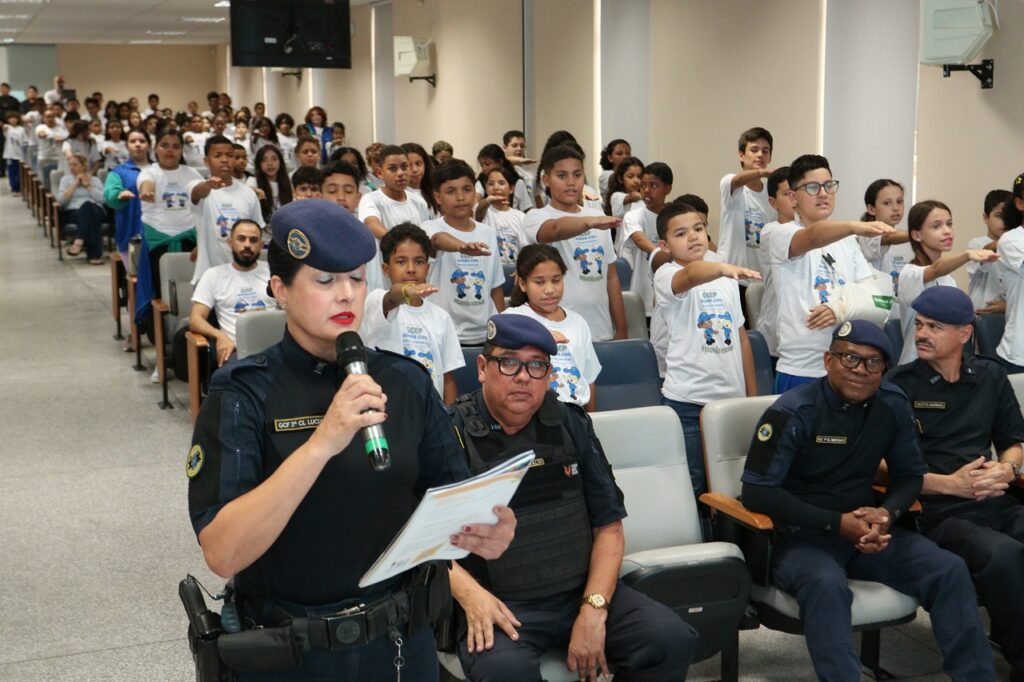 A imagem registra um momento de uma cerimônia de formatura do GCEP, realizada em um auditório. Ao centro, uma agente da Guarda Civil Municipal fala ao microfone enquanto segura documentos, acompanhada de outros integrantes da corporação.