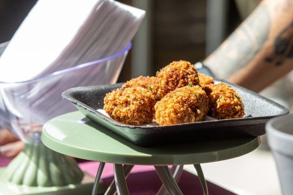 Prato de bolinhas empanadas e crocantes em close, servidas numa pequena travessa escura sobre um suporte verde. Comida: várias bolinhas douradas e uniformemente empanadas, com cobertura crocante e granulada; algumas pontinhas mais escuras indicam fritura bem dourada. Pequenos pedaços verdes (provavelmente salsinha) salpicados por cima. Apresentação: travessa retangular preta com papel por baixo das bolinhas; colocada sobre um suporte/pequena mesa verde fosca que aparece em primeiro plano. Iluminação e textura: luz natural intensa vindo da esquerda realça o brilho do empanado e cria sombras marcadas; textura crocante visível em detalhe. Fundo e contexto: ao fundo, elementos desfocados — um copo/recipiente transparente com guardanapos ou papéis brancos e, à direita, um braço com tatuagem parcialmente visível (desfocado), sugerindo ambiente casual de restaurante ou mesa ao ar livre. Atmosfera: imagem apetitosa e convidativa, foco nítido nas bolinhas que transmitem sensação de crocância e calor.