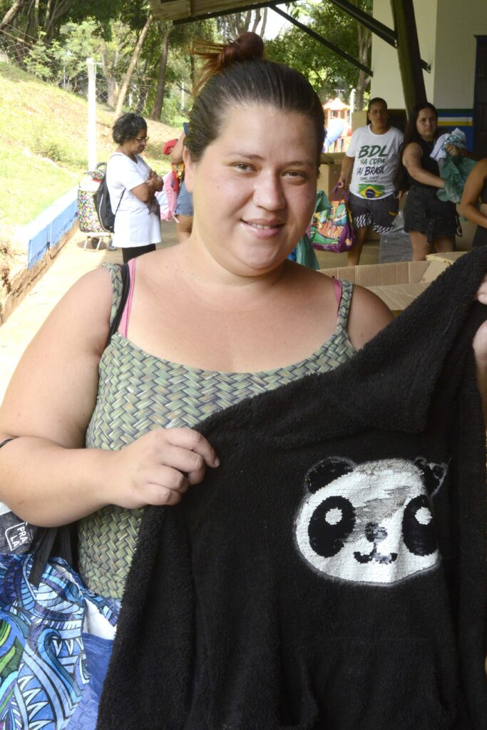 A imagem mostra uma mulher sorridente segurando uma blusa preta com o desenho de um panda bordado na frente. Ela está em um ambiente externo coberto, com outras pessoas ao fundo, algumas carregando sacolas e roupas, o que sugere uma ação comunitária ou campanha de doação. O cenário tem árvores e um muro pintado de azul, transmitindo um clima acolhedor e de solidariedade.