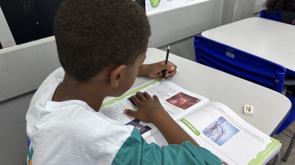 A imagem mostra um estudante sentado em uma carteira escolar, escrevendo com um lápis em um livro didático aberto. Ele tem cabelos curtos e cacheados, veste uma camiseta branca e está concentrado na atividade. Ao lado, há um estojo ou caixa azul sobre outra carteira. As páginas do livro apresentam textos, ilustrações e exercícios educativos, incluindo imagens relacionadas ao corpo humano e à saúde.